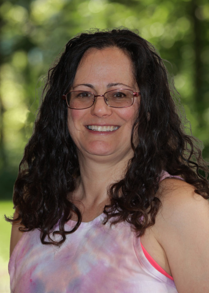 Heather, a white woman with long, dark, and curly hair stands in front of a blurred green background. She is wearing a pink tie-dye tank top, is smiling and has oval glasses. 