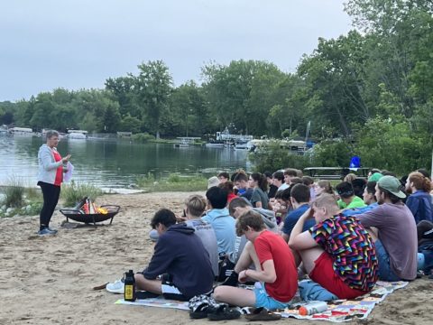 campers sit at beach listening to camp pastor at camp fire